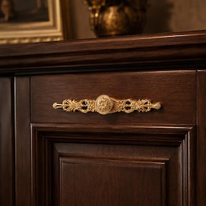 May include: Close-up of a dark brown wooden cabinet door with a decorative gold-colored handle. The handle features an ornate design with floral and circular patterns, adding a touch of elegance to the furniture.