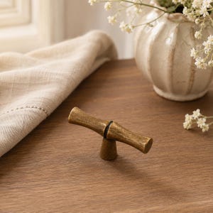 May include: A T-shaped cabinet knob with a textured, antique brass finish. The handle features a slightly curved, bamboo-inspired design with a central dark ring detail, supported by a single conical base that tapers towards the bottom.