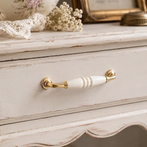 May include: A drawer handle featuring a central white ceramic bar with a ribbed texture, flanked by polished gold-tone metal mounts. The handle is attached to a distressed off-white wooden drawer front with visible wood grain and paint wear.