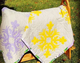 May include: A white quilt with a purple and yellow floral design. The quilt is folded and draped over a wooden chair.
