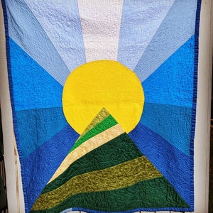 May include: A colorful quilt with a yellow sun and a green mountain design. The quilt is made of different shades of blue, green, and yellow fabric.