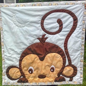 May include: A blue and white baby quilt with a brown monkey applique. The monkey has a curled tail and a brown face with yellow fur.