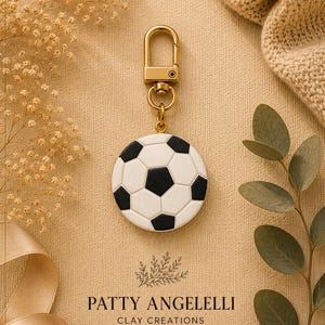 May include: A circular soccer ball charm featuring a classic black and white hexagonal pattern, attached to a gold-toned metal lobster clasp. The charm is presented with the text PATTY ANGELELLI CLAY CREATIONS HANDMADE WITH LOVE below it.
