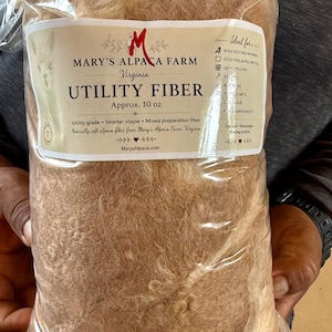 Pode incluir: Um saco plástico transparente cheio de fibra de alpaca crua de cor castanho-claro. O rótulo diz Mary's Alpaca Farm Virginia Utility Fiber, Approx. 10 oz, Utility grade, Shorter staple, Mixed preparation fiber, Naturally soft alpaca fiber.