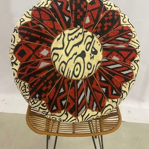 May include: Round, patterned floor cushion with a central medallion and a floral-like design. The cushion features red, black, and cream-colored geometric patterns. It rests on a brown woven stool with black metal legs, creating a decorative accent.