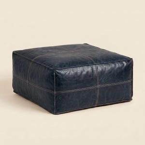 May include: A square, low-profile floor ottoman upholstered in dark navy blue leather. The surface features a grid of visible, light-coloured stitched seams that divide the top and sides into panels, creating a structured, geometric appearance.