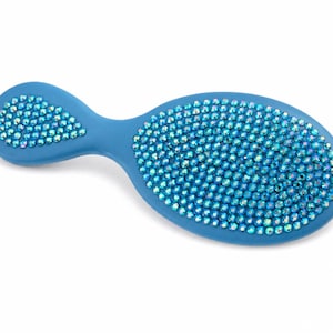 May include: A blue hairbrush with an oval head and a short handle. The back of the brush is densely covered in small, iridescent blue crystals that reflect light, creating a shimmering effect across the entire surface of the brush head and handle.