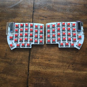 Peut inclure: Un kit de clavier mécanique ergonomique divisé composé de deux circuits imprimés blancs équipés de nombreux interrupteurs mécaniques rouges. Chaque moitié comprend un petit écran OLED rectangulaire monté dans le coin supérieur avec un couvercle transparent.
