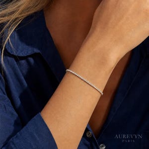 Pode incluir: Uma delicada pulseira de ténis com uma fila contínua de pequenas pedras transparentes de corte redondo, engastadas numa fina banda de metal prateado polido. A pulseira é usada no pulso, exibindo um design minimalista com um fecho discreto.