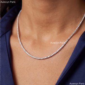 Puede incluir: Un collar de tenis en tono plateado que presenta una fila continua de pequeñas piedras redondas, claras y brillantes engastadas en una cadena metálica. El collar descansa sobre el cuello y el texto Aurevyn Paris es visible en la imagen.