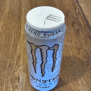 May include: A silver Monster Energy Ultra Zero Sugar can with a white lid. The can features the Monster logo in a light brown color and the words "ZERO SUGAR" and "ULTRA". The can is on a wooden surface.
