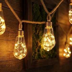 Op de afbeelding: Slingers met heldere, druppelvormige lampen, elk gevuld met warmwitte LED-lampjes. De lampen hangen aan een natuurlijk touw, wat een gezellige en uitnodigende sfeer creëert. De achtergrond is van hout en groen.