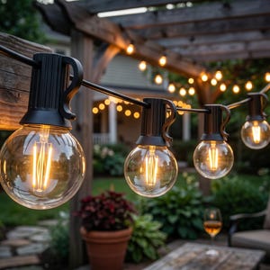 May include: A string of outdoor globe lights featuring clear glass bulbs with visible vertical filaments. The bulbs are attached to a black cord with integrated hanging hooks, emitting a warm glow against a dark evening outdoor setting.