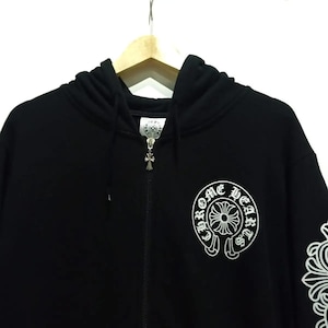 May include: Black zip-up hoodie featuring a white graphic on the chest and sleeve. The graphic includes the text "Chrome Hearts" in a circular design. The zipper pull and hood hardware are silver.