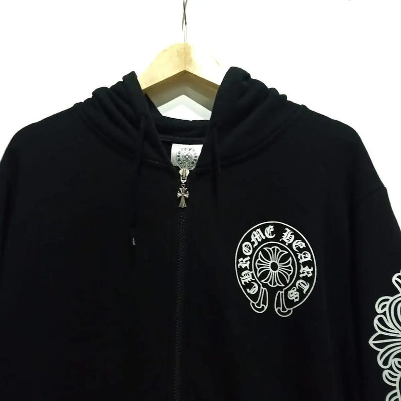May include: Black zip-up hoodie featuring a white graphic on the chest and sleeve. The graphic includes the text "Chrome Hearts" in a circular design. The zipper pull and hood hardware are silver.