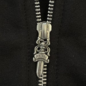 Może przedstawiać: Zbliżenie srebrnego suwaka zamka błyskawicznego z wygrawerowanym słowem "chrome". Zamek błyskawiczny znajduje się na czarnej tkaninie. Suwak ma ozdobny wz&oacute;r.