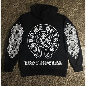 May include: Black hoodie with white graphic design. The back features a circular design with the words "Chrome Hearts" and a cross emblem. The sleeves have white decorative patterns. The text "LOS ANGELES" is printed below the circular design.