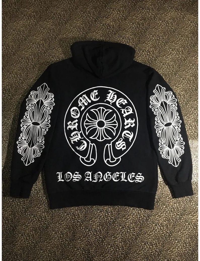 May include: Black hoodie with white graphic design. The back features a circular design with the words "Chrome Hearts" and a cross emblem. The sleeves have white decorative patterns. The text "LOS ANGELES" is printed below the circular design.