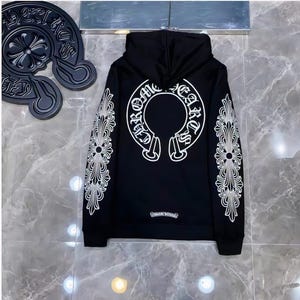 Może przedstawiać: Czarna bluza z kapturem z białymi, ozdobnymi wzorami na rękawach i plecach. Tył posiada duży biały emblemat ze słowami "Chrome Hearts" w okrągłym wzorze. Małe logo znajduje się na dole.