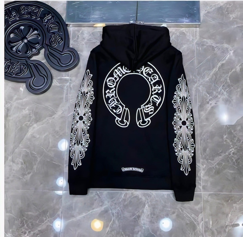 Może przedstawiać: Czarna bluza z kapturem z białymi, ozdobnymi wzorami na rękawach i plecach. Tył posiada duży biały emblemat ze słowami "Chrome Hearts" w okrągłym wzorze. Małe logo znajduje się na dole.