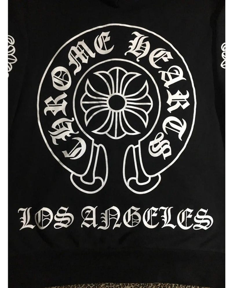 May include: Black sweatshirt with a large white graphic. The graphic features a circular design with the words "Chrome Hearts" and "Los Angeles" in a gothic font. A floral cross is in the centre of the circle.