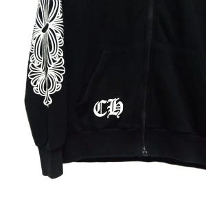 May include: Black zip-up hoodie featuring a white floral pattern on the left sleeve and the letters "CH" on the front. The hoodie has a front pocket and a zip closure.