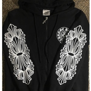 May include: Black zip-up hoodie with white ornate floral designs on the sleeves. The hoodie features a hood, a front zipper, and a small logo on the chest. The sleeves have large, symmetrical white patterns.