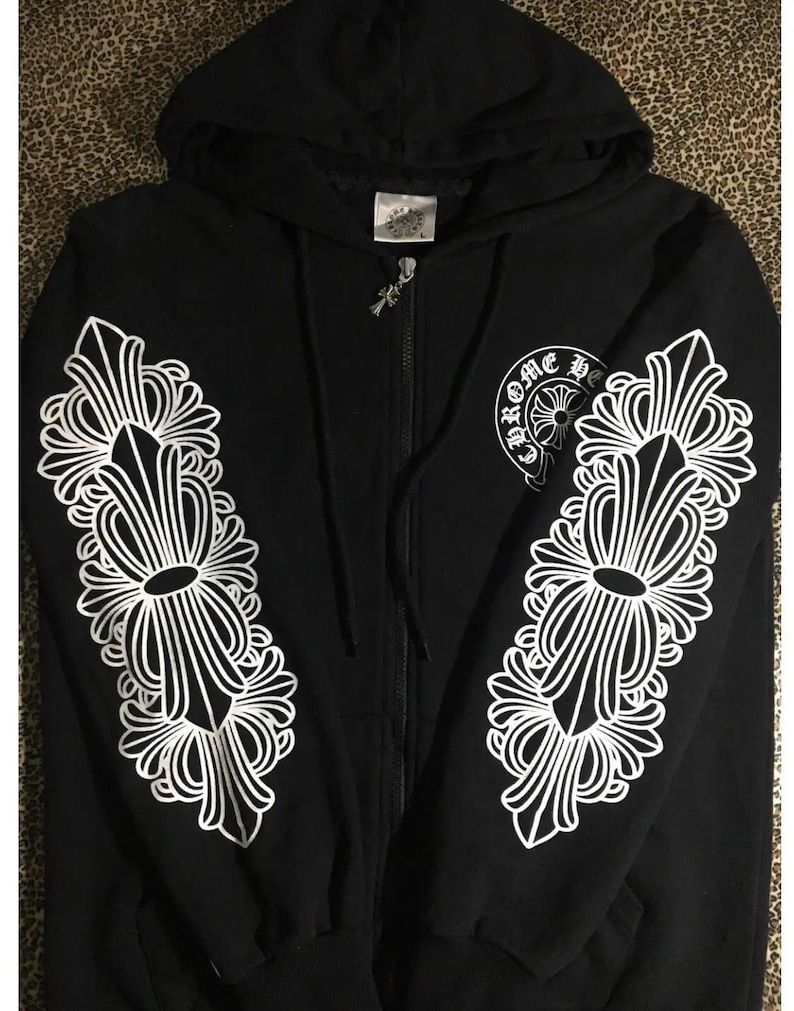 May include: Black zip-up hoodie with white ornate floral designs on the sleeves. The hoodie features a hood, a front zipper, and a small logo on the chest. The sleeves have large, symmetrical white patterns.