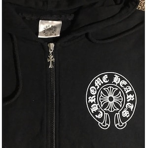 May include: Black zip-up hoodie with a hood and a silver zipper pull. The hoodie features a white graphic of a circular design with the words "Chrome Hearts" around it. A tag with a logo is visible near the hood.