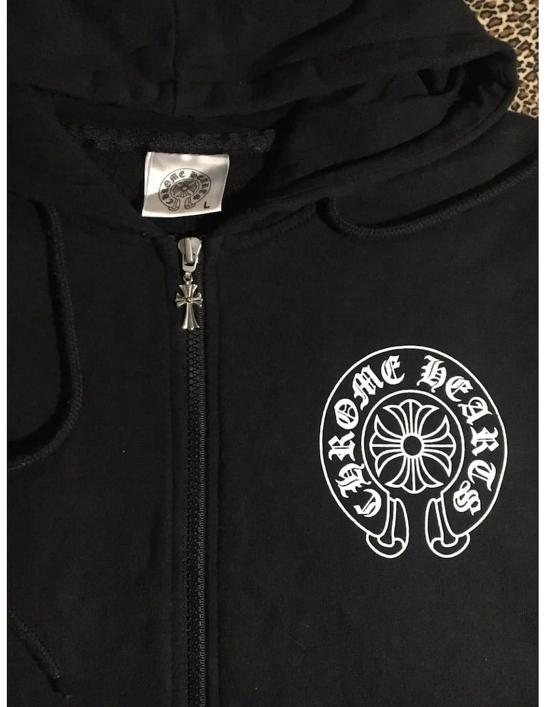 May include: Black zip-up hoodie with a hood and a silver zipper pull. The hoodie features a white graphic of a circular design with the words "Chrome Hearts" around it. A tag with a logo is visible near the hood.