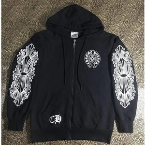 May include: Black zip-up hoodie with white ornate designs on the sleeves and a logo on the chest. Features a hood and front pockets. The hoodie has a silver zipper and the letters "CH" on the front.