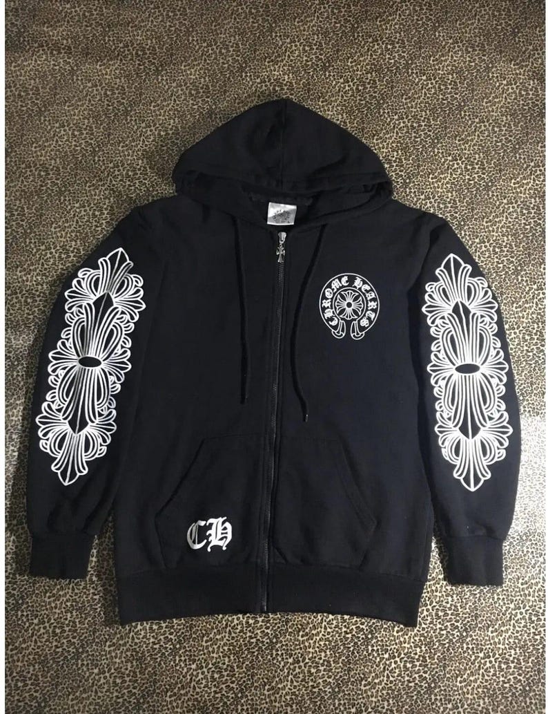 May include: Black zip-up hoodie with white ornate designs on the sleeves and a logo on the chest. Features a hood and front pockets. The hoodie has a silver zipper and the letters "CH" on the front.