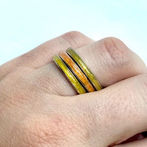 May include: A set of three thin metal rings worn together, each featuring a smooth, inlaid band of glittery resin in shades of bright yellow, vibrant orange, and lime green, creating a stacked appearance with a polished metallic finish.