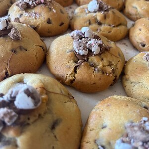 May include: Close-up of several freshly baked chocolate chip cookies. The cookies are golden brown with visible chocolate chips and are topped with a cluster of chocolate and candy pieces. The cookies are arranged on a light-colored surface.