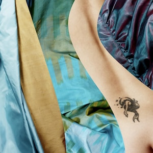 Könnte beinhalten: Ein Arm mit einem schwarzen Meerjungfrau-Tattoo neben einer Sammlung farbenfroher Stoffe. Die Stoffe umfassen Hellblau, Gold und Türkis mit Streifenmustern. Die Stoffe scheinen Seide oder ein ähnliches Material zu sein.