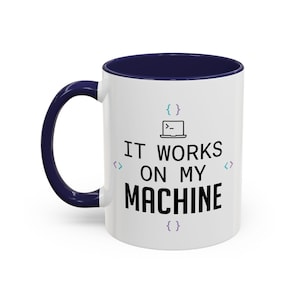 Puede incluir: Una taza de cerámica blanca con interior y asa de color azul oscuro. El frente presenta un gráfico negro de una computadora portátil con un icono de símbolo del sistema, el texto IT WORKS ON MY MACHINE y cuatro juegos de corchetes coloridos.