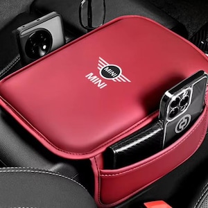 Könnte beinhalten: Eine rote Armlehnenabdeckung für die Mittelkonsole mit dem weißen MINI-Logo. Dieses rechteckige Zubehörteil verfügt über eine seitliche Aufbewahrungstasche mit einem schwarzen Smartphone und einer schwarzen Geldbörse für die Armlehne.