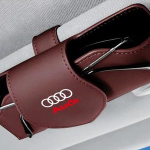 Puede incluir: Un estuche para gafas de coche en cuero marrón oscuro con el logotipo de Audi en blanco y rojo en la solapa frontal. El estuche está sujeto al parasol de un vehículo y contiene unas gafas con montura plateada visibles en el compartimento.