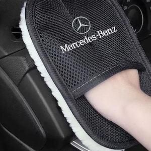 Op de afbeelding: Een zwarte autopoetshandschoen met een witte pluche voering en een mesh buitenkant. De handschoen is voorzien van het Mercedes-Benz logo en tekst in wit borduurwerk op de bovenzijde. Het ontwerp heeft een vak om de hand in te steken.