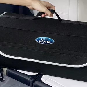 May include: A black rectangular car boot organiser featuring a fabric handle on top and a blue and white Ford logo centred on the front flap. The organiser has a light grey trim along the edges of the flap and is constructed from a felt-like material.