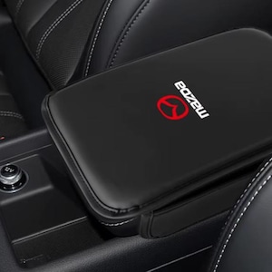 Puede incluir: Una funda de cuero negro para el reposabrazos de la consola central con el logotipo de Mazda en rojo y blanco. La funda rectangular está colocada sobre el compartimento de la consola central, con una toma de corriente de 12V visible cerca.