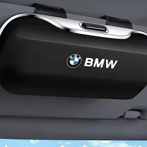 May include: A black rectangular car visor sunglasses holder with a silver metallic trim along the top edge. The front surface features the white and blue BMW logo followed by the white text BMW printed in a clean, sans-serif font style.