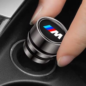 May include: A cylindrical metallic car accessory with a knurled texture and a black top surface featuring the letter M in white, accompanied by three diagonal stripes in light blue, dark blue, and red, designed to fit into a vehicle power outlet.