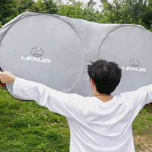 Puede incluir: Un parasol plateado y plegable para el parabrisas del coche, con dos paneles circulares que muestran el logotipo y el texto de Lexus impresos en blanco en cada lado. El parasol tiene un borde de tela negra para bloquear la luz solar.