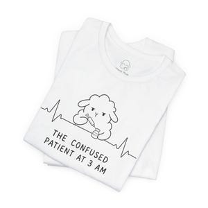 Può includere: Due magliette bianche sovrapposte, quella superiore con una grafica nera di una pecora che tiene una siringa e una fiala. Il testo recita "THE CONFUSED PATIENT AT 3 AM" con una linea del battito cardiaco. Il logo del marchio "Sheep Mode" è visibile.