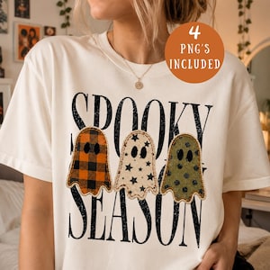 May include: A cream-coloured t-shirt featuring the black text "SPOOKY SEASON" and three ghost illustrations. The first ghost has an orange and black buffalo plaid pattern, the second is cream with black stars, and the third is olive green with black polka dots.