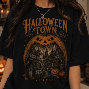Puede incluir: Una camiseta negra con un gran estampado gráfico naranja con el texto HALLOWEEN TOWN en la parte superior y EST. 1998 en la parte inferior. El diseño muestra una escena de pueblo espeluznante dentro de una calabaza gigante, con un gato negro.