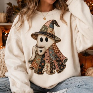 May include: A cream-colored crewneck sweatshirt featuring a large graphic of a ghost wearing a patchwork-style witch hat and cloak. The ghost holds a small beige mug with a black heart icon. The patchwork design includes various autumn patterns.