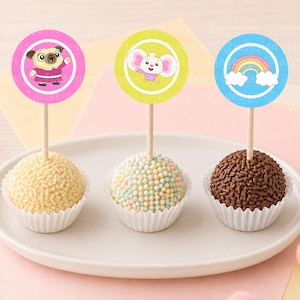 Pode incluir: Três cake pops em um prato branco, cada um com um topper redondo decorativo. Os toppers apresentam um pug, um elefante e um arco-íris. Os cake pops são decorados com granulado e chocolate.
