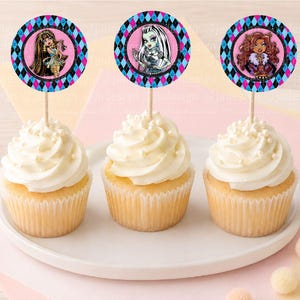 Pode incluir: Três cupcakes com topos de papel redondos apresentando ilustrações de personagens com cabelos longos, pele verde clara e cabelos castanhos. Cada topo tem uma borda com padrão de losangos azul, preto e rosa em um palito de madeira.